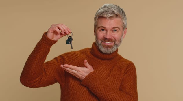 Middle-aged old man real estate agent lifting hand showing the keys of new home house apartment, buying or renting property, mortgage loan. Senior mature guy isolated alone on beige studio background