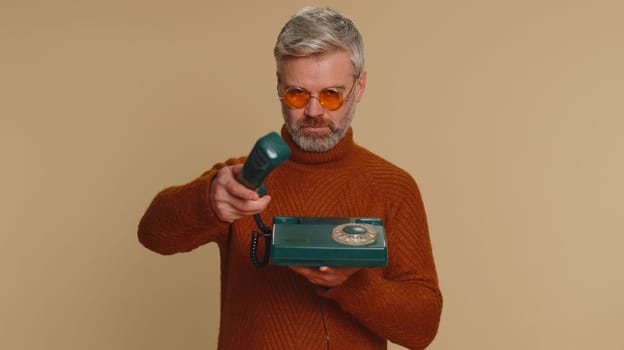 Hey you, call me back. Middle-aged old man talking on wired vintage old-fashioned telephone of 80s, advertising says hey you call me back. Senior mature guy isolated alone on beige studio background