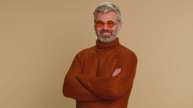 Seductive happy middle-aged old man wearing sunglasses, charming smile, celebrating success, winning, flirting. Positive expression. Senior smiling mature guy tourist alone on beige studio background