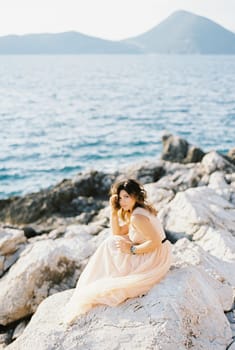 Bride sits on the rocks by the sea with her head propped up by her hand. High quality photo