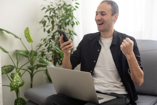 Smiling young man typing on mobile phone while sitting on a sofa at home with laptop computer.