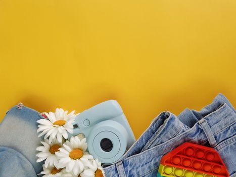 Summer bright flatlay on a yellow background. On a yellow background there is an antistress, a camera, a bouquet of daisies, jeans and a cap.