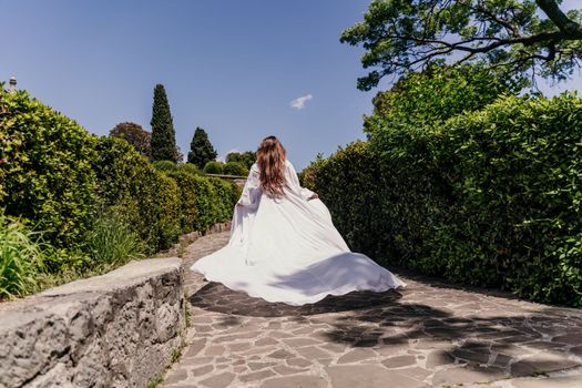 Brunette runs white dress park. A beautiful woman with long brown hair and a long white dress runs along the path along the beautiful bushes in the park, rear view.