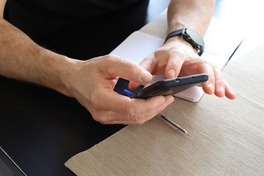 Close-up of the hands of a man with a smart watch on his hand, who is holding a black phone, looking for something. Black table with notepad and pen. Remote work concept.