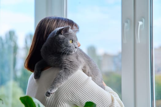 Woman hugging purring grey cat, holding pet in her arms, looking out the window together. Love, friendship, lifestyle, animals people concept