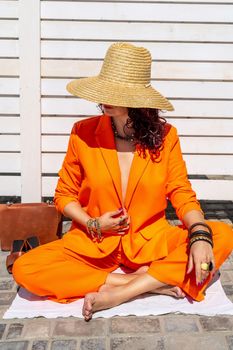 Stylish woman in an orange suit with a hat sits on a rug on a white striped background. On the hands are jewelry rings and bracelets, sandals and a bag stand side by side