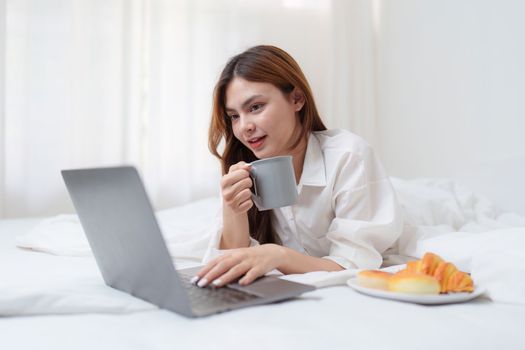 Beautiful asian woman on a white bed with coffee and using laptop computer at home. lifestyle concept