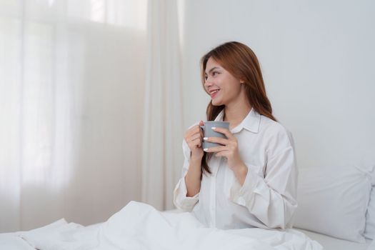 Beautiful asian woman has woken up on a white bed and holding coffee in the morning at home. lifestyle concept