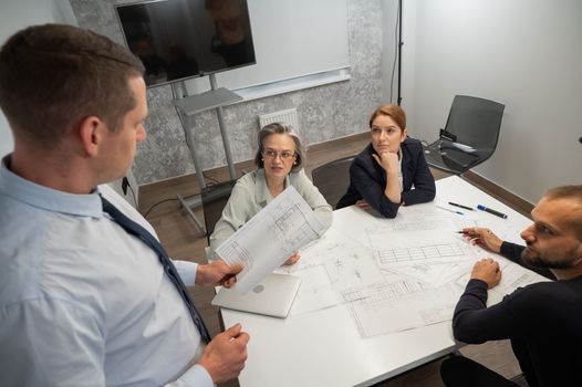 A caucasian man stands and holds a drawing, three colleagues sit at a table and listen to him