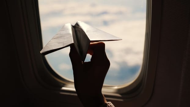 close-up. Silhouette of a child's hand with small paper plane against the background of airplane window. Child sitting by aircraft window and playing with little paper plane. during flight on airplane. High quality photo