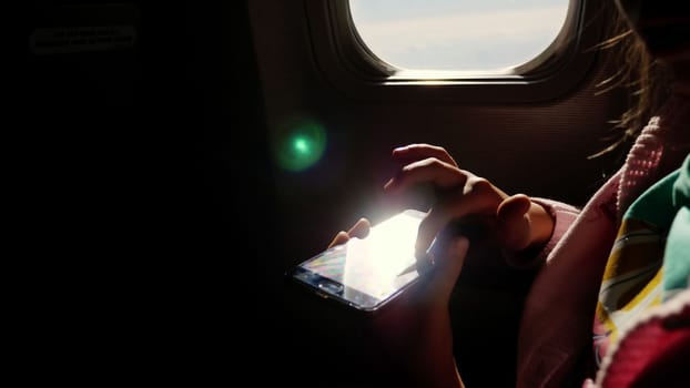 close-up. the sun's rays are reflected in the phone. dark silhouette of kid hands and mobile phone against airplane's illuminator. Child using, plays game on smartphone, tablet phone in airplane cabin. High quality photo