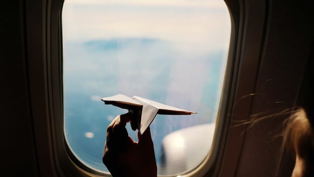 close-up. Silhouette of a child's hand with small paper plane against the background of airplane window. Child sitting by aircraft window and playing with little paper plane. during flight on airplane. High quality photo