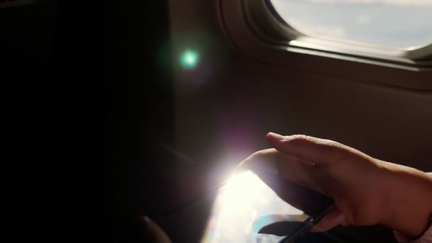 close-up. the sun's rays are reflected in the phone. dark silhouette of kid hands and mobile phone against airplane's illuminator. Child using, plays game on smartphone, tablet phone in airplane cabin. High quality photo