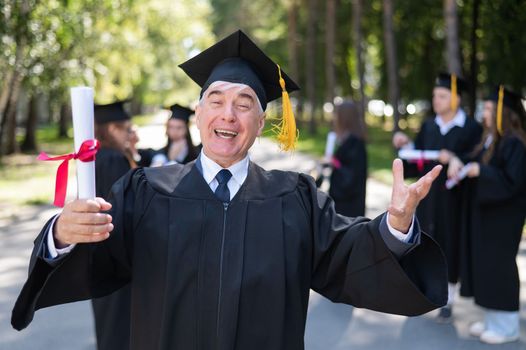 A group of graduates in robes outdoors. An elderly student rejoices at receiving a diploma