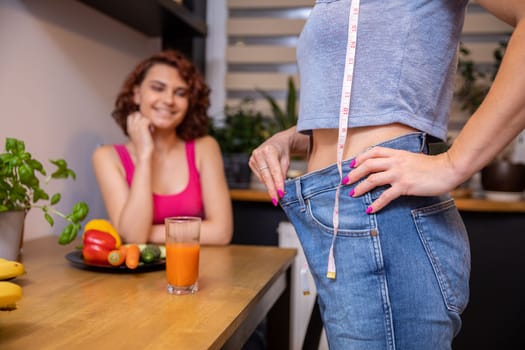 A friend sits at the kitchen table in the blurry background. The woman in the foreground stands sideways and pulls back the waistband of her too loose pants showing off the effects of weight loss.