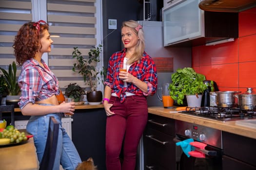 Women dressed in pin-up style stand in the kitchen and hold glasses of juice. Herbs in a pot stand on the kitchen counter. There are metal pots on the stove. The women are dressed in checkered shirts.