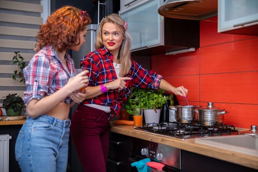 Two girls standing by a gas stove. The kitchen tiles are orange in color. The friends are standing close to each other and talking. One of them is stirring with a spoon in a metal pot standing on the gas.