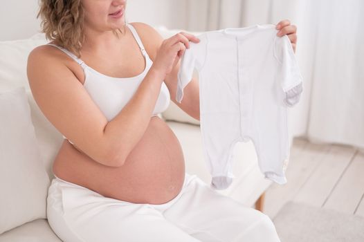 A pregnant woman sits on a white sofa and holds clothes for the expected baby