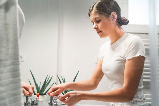 Young caucasian woman getting ready in a bright white modern bathroom. Woman doing her skincare, cleaning her face, teeth and body int he bathroom.