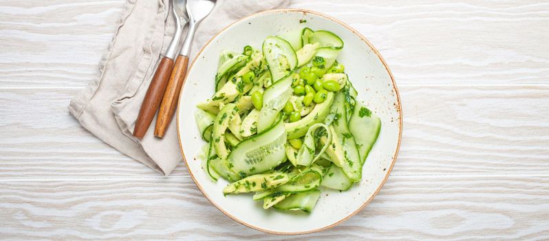 Healthy vegan green avocado salad bowl with sliced cucumbers, edamame beans, olive oil and herbs on ceramic plate top view on white wooden rustic table background