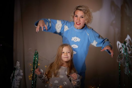 Cute mother and daughter in pajamas having fun in the room with Christmas garlands and white background. The tradition of decorating the house for holidays. Happy childhood and motherhood