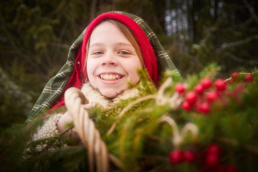 Nice cute teenager girl in peasant clothes with hot fur coat and shawl in cold winter forest with fir trees, pines and snow on winter day. Child with basket full of green branches and red berries