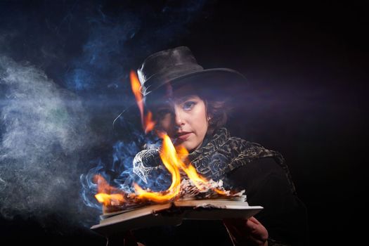 Portrain of witch girl with fier on a magic Book. Woman in hat conjuring, making witchcraft over spooky dark black magic background. Model posing in Halloween party. Author writer burns manuscript