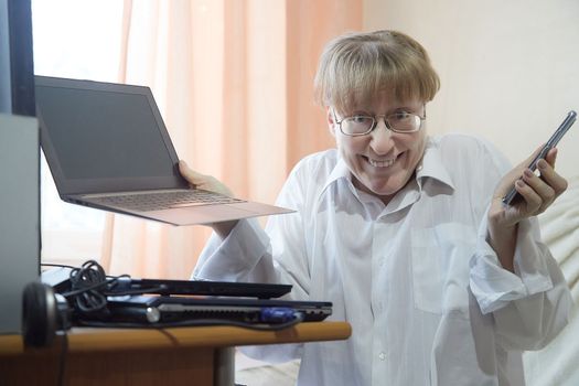 Beautiful slim blond woman in glass and white shirt with laptop computer and cell phone at home near window. Emotions from online work on Internet or chats. Joy, surprise, indignation. Partial focus