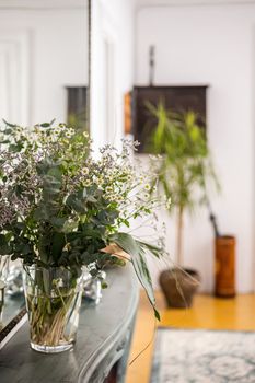 Close-up glass vase with bouquet of wildflowers stands on backdrop of a blurred stylish mediterranean interior in cozy apartment with plant. Decor and inspirational design concept.