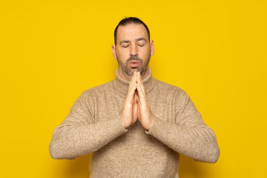 Bearded hispanic man wearing beige turtleneck isolated over yellow background, holding hands in prayer or meditation, looking relaxed and calm, dreaming and hoping for the best