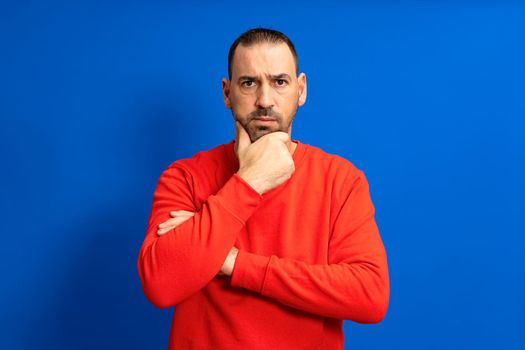 Bearded hispanic man wearing red sweater pensive thinking looking at copyspace hold hand chin isolated over blue color background