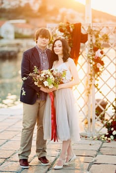 Groom and bride with a bouquet of flowers stand near the wedding screen on the pier. High quality photo