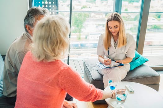 Young blonde woman caregiver helping senior married couple at home. Nurse assisting her senior patients at nursing home. Senior couple being helped by nurse at home. High quality photo