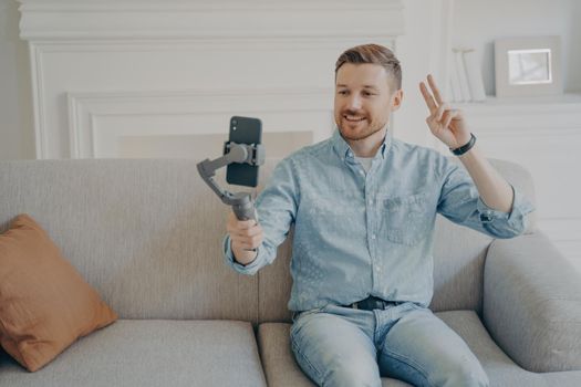 Young man with short beard wearing casual clothes and sitting on comfortable couch, video chatting holding gimbal with phone attached to it, showing peace gesture to camera