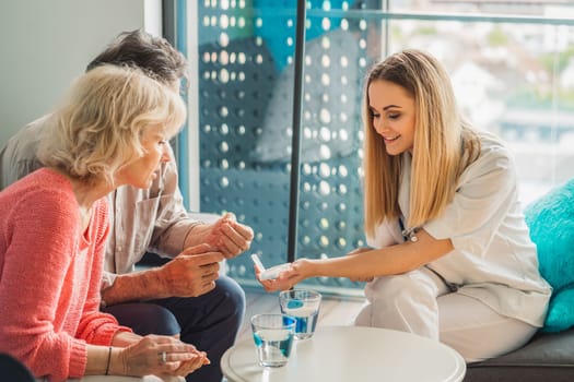 Young blonde woman caregiver helping senior married couple at home. Nurse assisting her senior patients at nursing home. Senior couple being helped by nurse at home. High quality photo