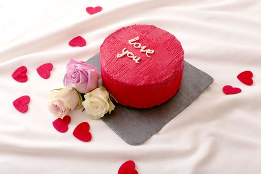 Small round red bento cake with the inscription Love you, with red hearts and roses lying on a white bed next to it . Close up. Copy space