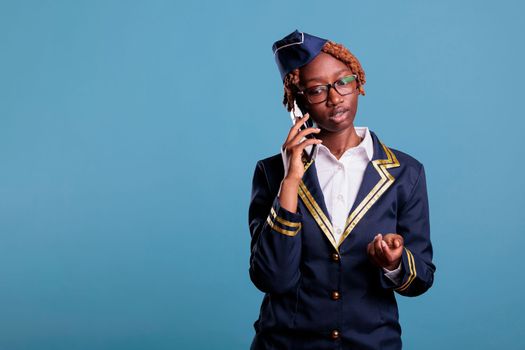 Concentrated female flight attendant talking on the phone dressed in uniform and eyeglasses. Stewardess looks serious while using mobile device in studio shot against blue background.