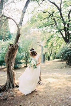 Bride with a bouquet stands near a tree in the park and looks at the hem of her dress. High quality photo
