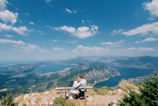 Bride and groom sit hugging on a bench on the mountain and look at the valley. High quality photo