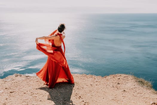 Side view a Young beautiful sensual woman in a red long dress posing on a rock high above the sea during sunrise. Girl on the nature on blue sky background. Fashion photo.