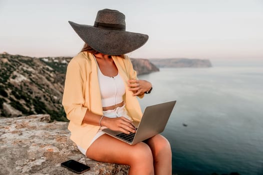 Successful business woman in yellow hat working on laptop by the sea. Pretty lady typing on computer at summer day outdoors. Freelance, travel and holidays concept.