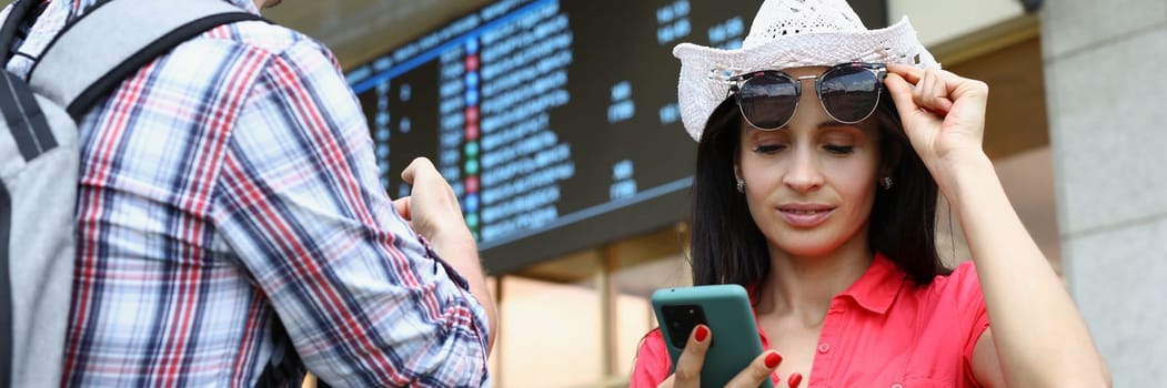 Man and woman are tourists with mobile phone against background of electronic board of bus and railway transport timetables. Smartphone airport transport concept app and flight schedules