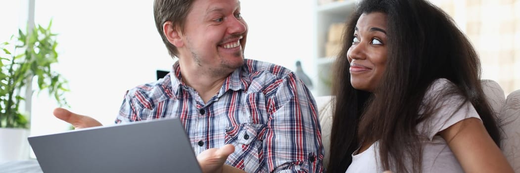 Interethnic family couple laughing and chatting while sitting on sofa at home with laptop. Married couple making online payments on laptop using financial banking app on computer at home