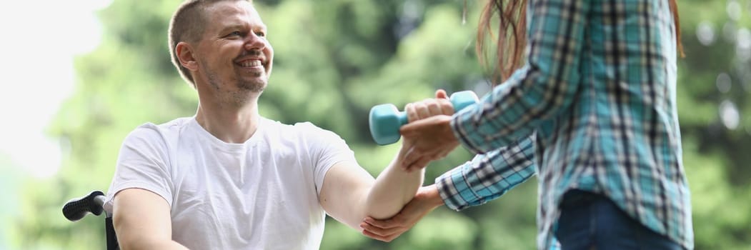 Female physiotherapist helps young man in wheelchair to lift weight of arms in park. Rehabilitation of disabled and physical activity