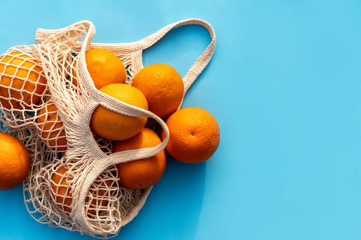 Mesh white cotton reusable bag with oranges on a blue background. Environmentally friendly shopping, the environment and the concept of healthy foods, citrus fruits with vitamin C. Place for text.