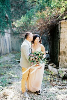 Groom hugs bride with a bouquet near the brick wall in the garden. High quality photo