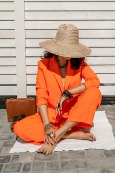 Stylish woman in an orange suit with a hat sits on a rug on a white striped background. On the hands are jewelry rings and bracelets, sandals and a bag stand side by side