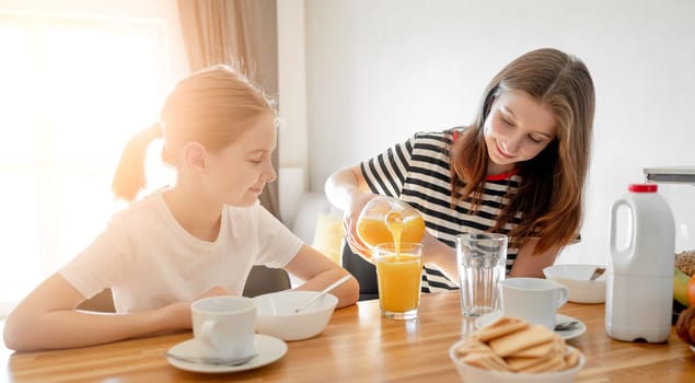 Girls sisters eat tasty breakfast together and filling glass with orange juice. Female child kid and subling with citrus beverage during morning meal