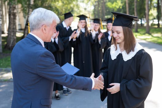 The teacher shakes hands with the student and presents the diploma outdoors. A group of university graduates