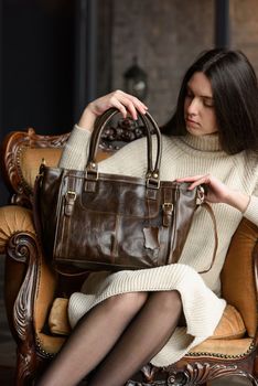 a brunette girl in a knitted beige dress poses while sitting with a shiny leather bag in her hands